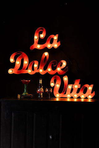 The La Dolce Vita Fairground Wall Light styled on the wall switched on, above a red flower vase, a bottle and some wine glasses.