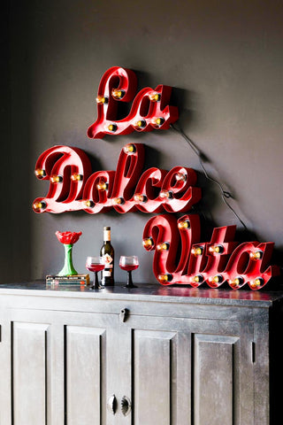 The La Dolce Vita Fairground Wall Light styled on a wall in daylight, above a sideboard with a flower vase, some books, a bottle and some wine glasses.