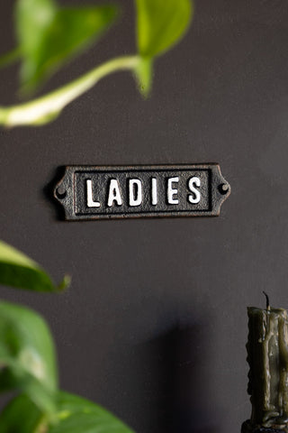 The Ladies Door Hanging Sign displayed on a black wall, styled with greenery and a green candle with dried wax running down it.