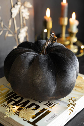 The Large Black Velvet Pumpkin displayed on a book on a dark surface with some fairy lights, with lit candles in the background.