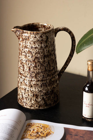 The Large Mottled Brown Jug on a black table with a book, bottle and a plant.