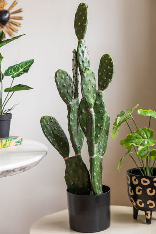 A large faux cactus plant in a black pot against a white wall. 