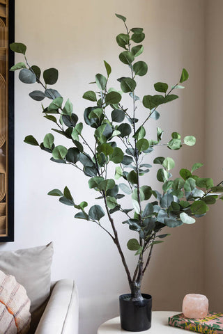 A lifestyle image of a large potted faux eucalyptus plant against a white wall. 