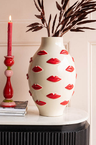 The Large Red Kiss Me Lips Flower Vase on a sideboard, styled with bamboo stems inside, next to a lit candle and some magazines.