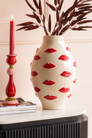 The Large Red Kiss Me Lips Flower Vase styled with a bamboo stem inside, on a sideboard with a lit candle and some magazines.