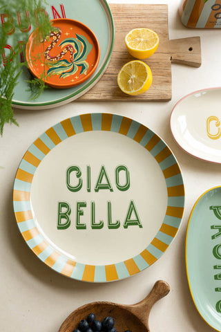 The Large White Ciao Bella Plate styled on a colourful tablescape of plates, fruit, kitchen accessories and a plant.