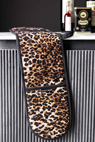 The Luxury Leopard Print Oven Gloves displayed on the edge of a table with some cooking accessories in the background.