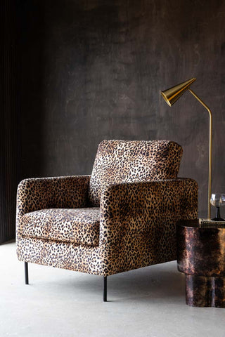 The Edie Leopard Print Armchair styled in front of a dark wooden wall with a side table, floor lamp, book and glass.