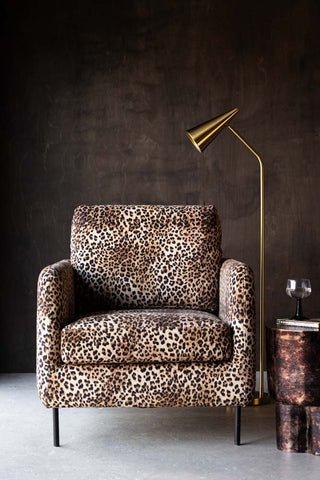 The Edie Leopard Print Armchair seen from the front, styled in front of a dark wooden wall with a gold floor lamp, side table, book and wine glass.