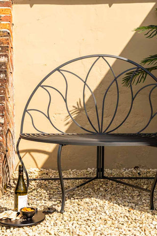 The Lotus Leaf Rounded Back Garden Bench in a garden in the sun in front of a wall, styled with a plant, a wine bottle and serving board with food.