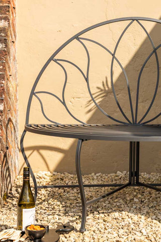 Close-up shot of the Lotus Leaf Rounded Back Garden Bench in front of a wall in the sun, style with a wine bottle, serving board and food.