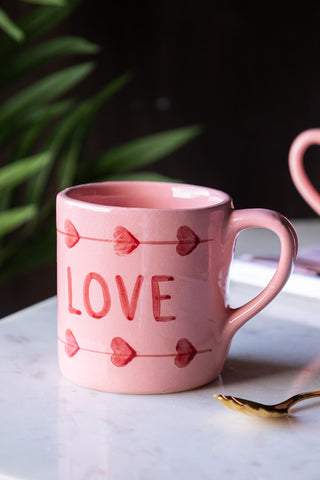 The Love Hand-Painted Cappuccino Mug on a white surface with a spoon, magazine, mug and plant in the background.