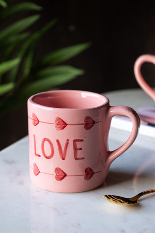 The Love Hand-Painted Cappuccino Mug displayed on a white marble surface with other kitchen accessories and a plant in the background.