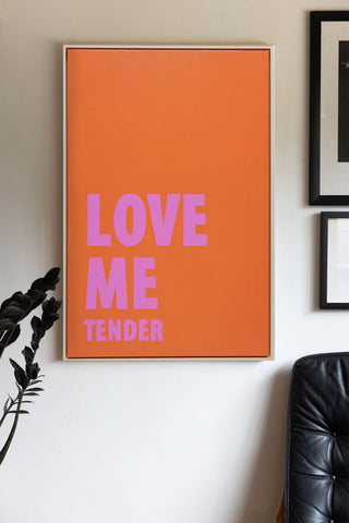 The Love Me Tender Canvas displayed on a white wall next to some art prints, a plant and a chair.