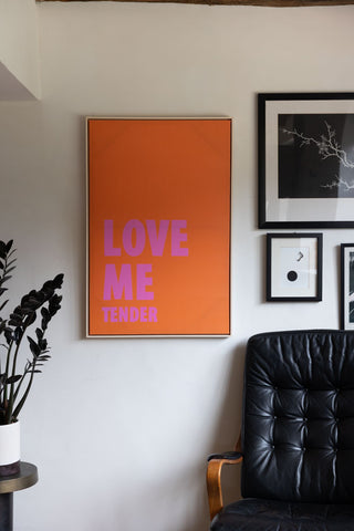 The Love Me Tender Canvas styled on a white wall next to other art prints, with a chair, table and plant.