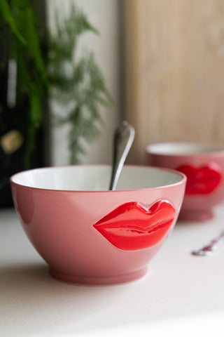 Pink ceramic cereal bowl with a 3D red lips design, filled with a silver spoon, placed on a light surface.