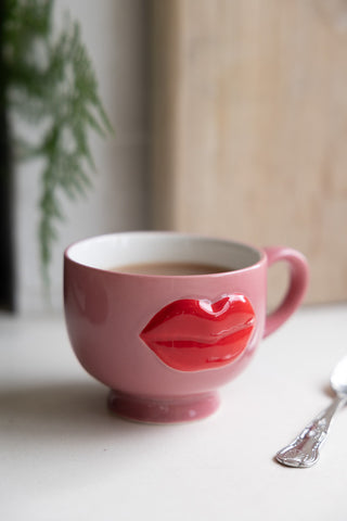 Charming pink mug with raised red lips detail, filled with a warm beverage and placed next to a vintage-style spoon