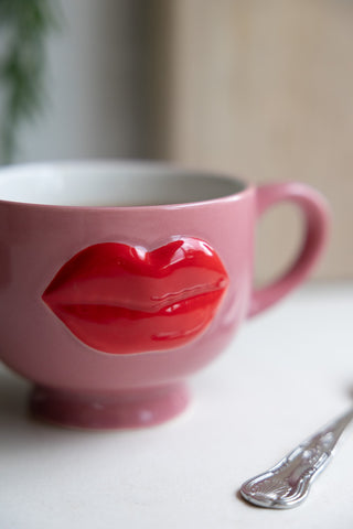 Cute pink mug with a glossy 3D red lips design, filled with tea, placed on a white table beside a decorative spoon.