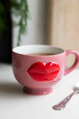 Pink ceramic mug with a 3D design of red lips, filled with tea on a table next to a silver spoon.