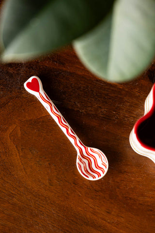 The Made With Love Red Striped Spoon styled on a wooden surface with a mug and plant.