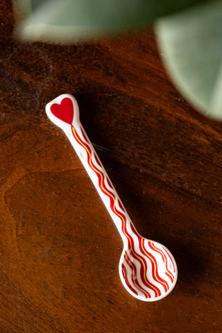 The Made With Love Red Striped Spoon styled on a wooden surface, with the leaves of a plant at the top of the shot.