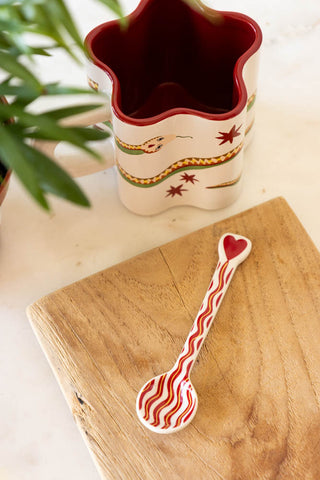 The Made With Love Red Striped Spoon displayed on a wooden chopping board next to a mug and plant.