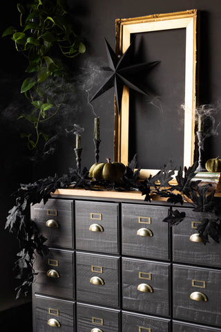 The Maple Leaf Garland styled across a black sideboard with some candles that have just been extinguished, Halloween decorations, a large gold frame and some greenery.