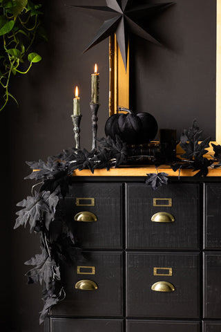 The Maple Leaf Garland styled on a black and wooden sideboard, with some lit candles, Halloween decorations, gold frame and some greenery.