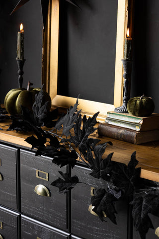 Close-up of the Maple Leaf Garland draped across a black and wooden sideboard, styled with some lit candles, Halloween decorations, a frame and some books.