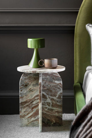 The White Marble Round Side Table styled with a small table lamp and a mug on, displayed next to a bed as a bedside table.