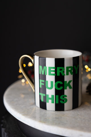 The Merry Fuck This Stripe Mug styled on a white surface with fairy lights in the background.