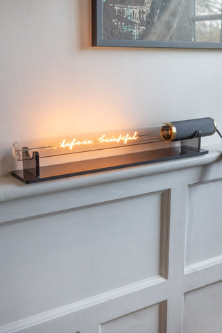 The Message In A Tube Light - Life Is Beautiful on the stand, switched on and displayed on a shelf in front of a wall beneath an art print.