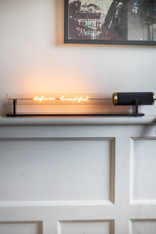 The Message In A Tube Light - Life Is Beautiful switched on and displayed on a shelf, in front of a white wall with a framed art print on.