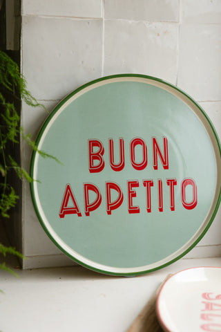 The Mint Green Buon Apetito Plate styled leaning against a tiled kitchen wall next to another plate and plant.