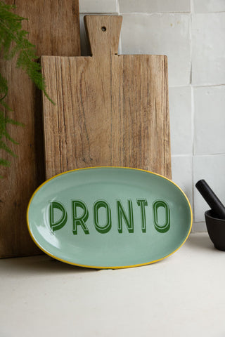 The Mint Green Pronto Plate styled in a kitchen leaning against two wooden chopping boards, next to a pestle and mortar and plant.