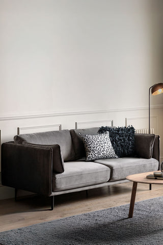 The Modern Charcoal Grey Velvet 3-Seater Sofa styled with some cushions on, displayed in a living room behind a coffee table on a rug, with a floor lamp in the background.