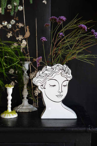 The Peaceful Monochrome Lady Vase styled with flowers inside on a sideboard next to some candlestick holders and greenery.