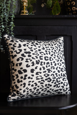 An image showing the back of The Monochrome Leopard Sit The Fuck Down Cushion styled on a dark bench, with a sideboard styled with various accessories in the background.