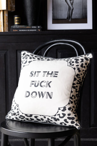 The Monochrome Leopard Sit The Fuck Down Cushion styled on a black dining chair, with a sideboard styled with various accessories in the background.