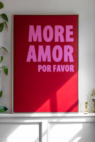 The More Amor Canvas displayed leaning against a white wall, next to a plant, candlestick holder and ornament.