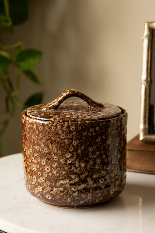 The Mottled Brown Lidded Pot styled on a white surface, with some home accessories and a plant in the background.