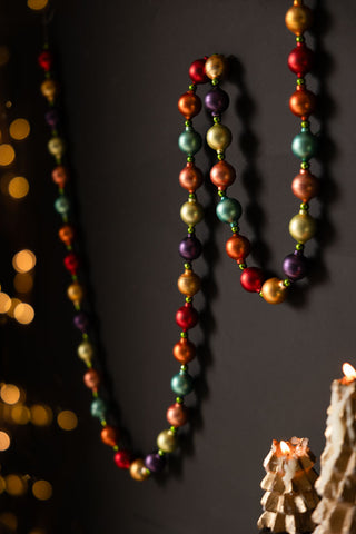 The Multi-Colour Bauble Garland displayed draped on a black wall, with fairy lights and some lit candles also in the shot.