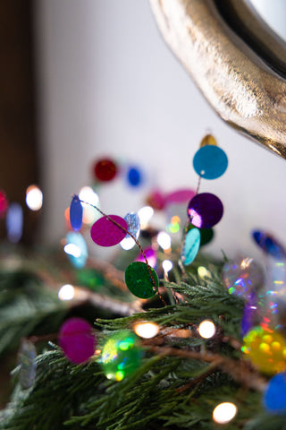 A close up of The Multi Coloured Confetti Decorative Light Chain styled on a mantlepiece with greenery