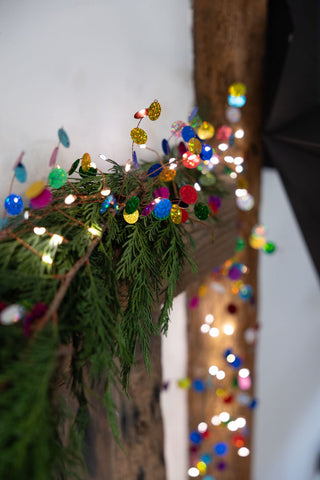 A wide shot of The Multi Coloured Confetti Decorative Light Chain styled on a mantlepiece with greenery