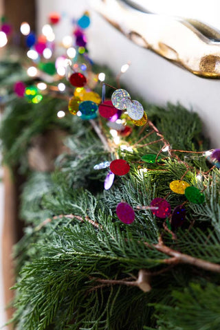 The Multi Coloured Confetti Decorative Light Chain styled on a mantlepiece with greenery and a mirror behind