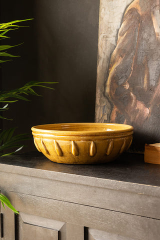 The Mustard Ceramic Serving/Fruit Bowl on a sideboard with a canvas and book, with a plant to the side of the shot.