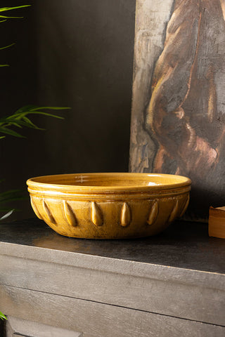 The Mustard Ceramic Serving/Fruit Bowl displayed on a black surface with a canvas, book and a plant.