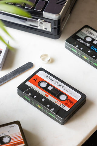 The My Favourite Tunes Cassette Tape Storage Tin styled on a white marble surface with some other tins, a plant and some small accessories.