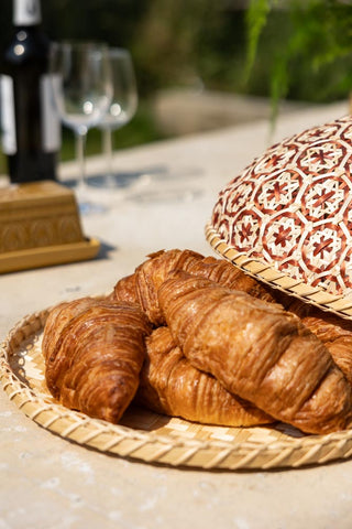 The Natural Bamboo & Red Floral Detail Food Cover styled on a table with the lid leaning on some croissants.