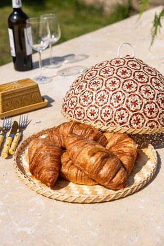 The Natural Bamboo & Red Floral Detail Food Cover styled on an outdoor table, with croissants and various tableware.